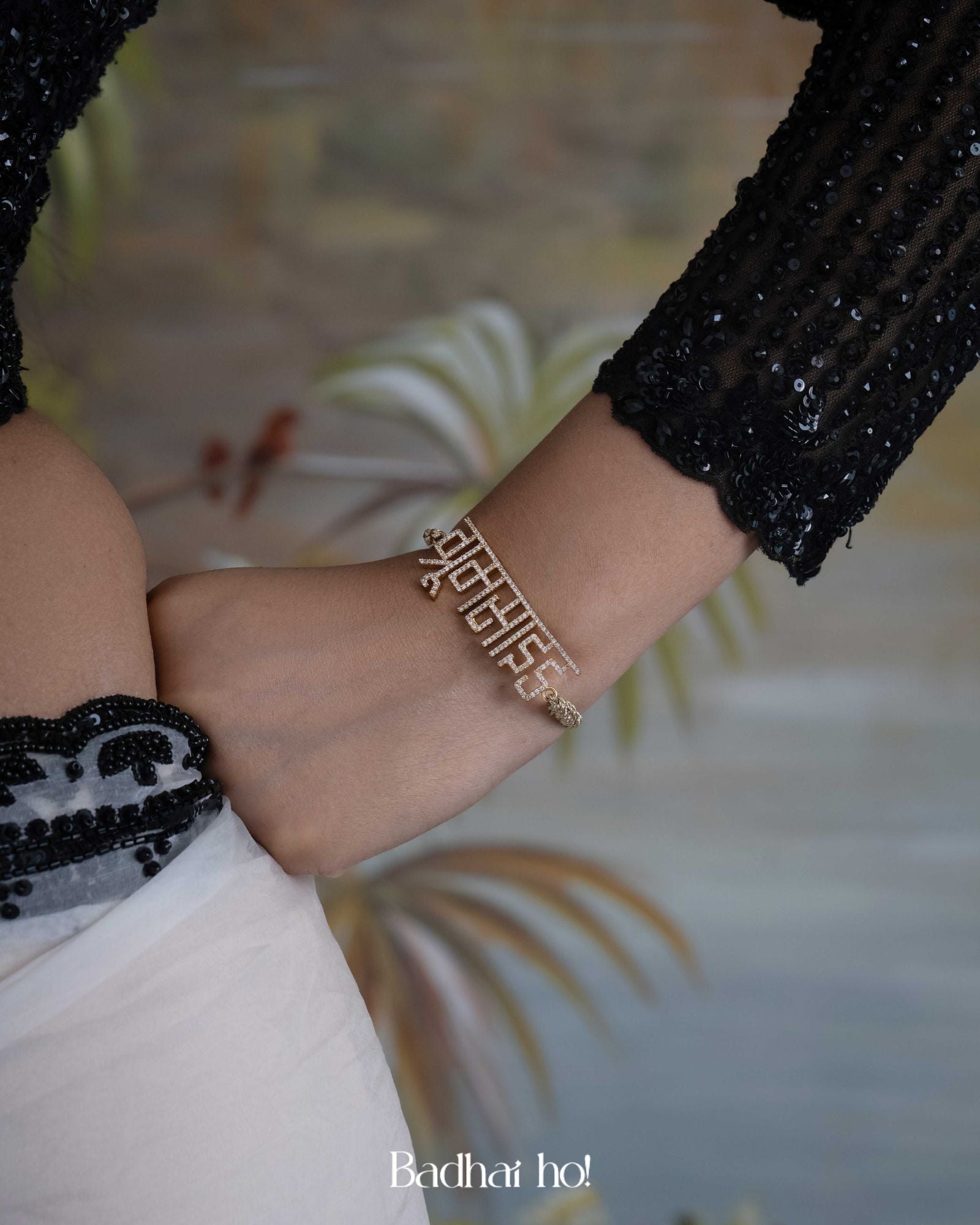 Close-up of a wrist wearing a gold bracelet with text, against a blurred indoor background.