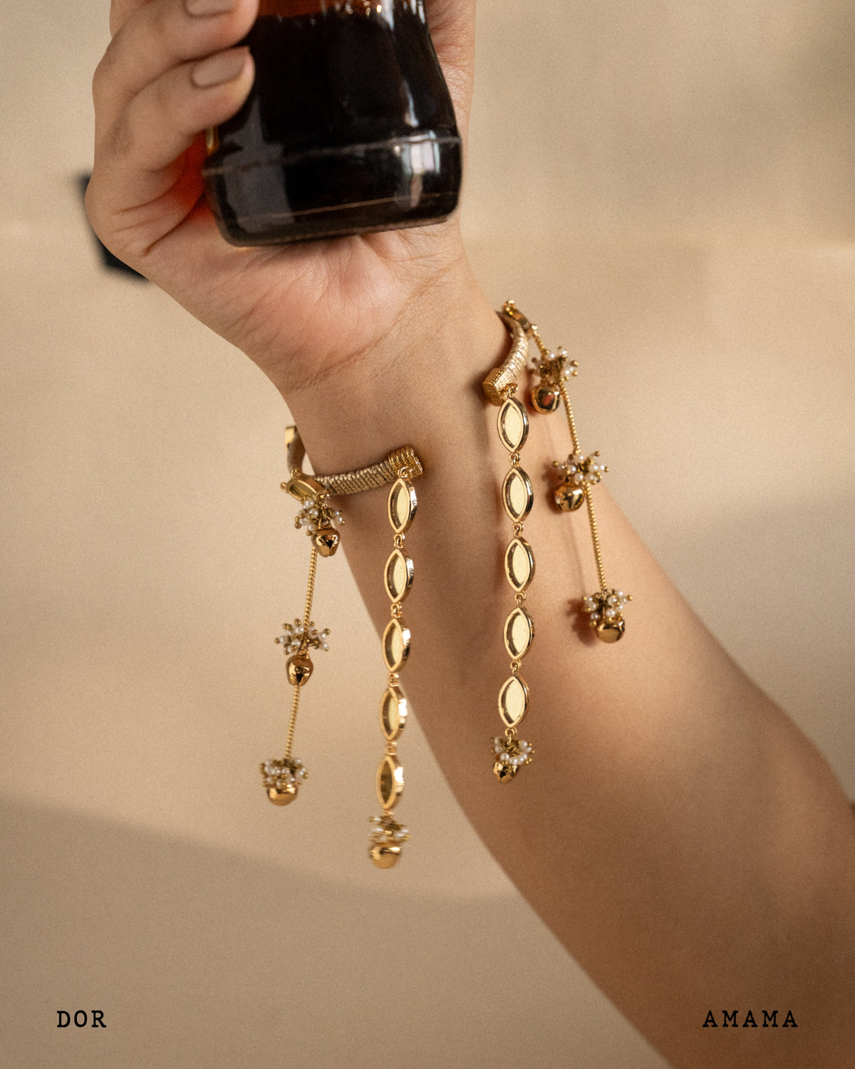 Gold rakhi lumba being held by a hand against a beige background, with 'DOR' and 'AMAMA' branding visible.

