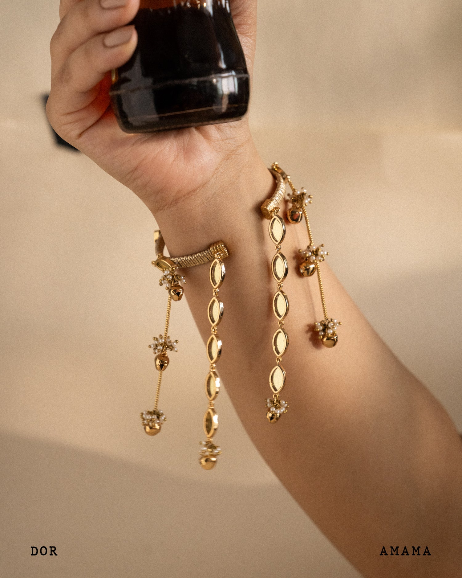 Gold rakhi lumba being held by a hand against a beige background, with 'DOR' and 'AMAMA' branding visible.

