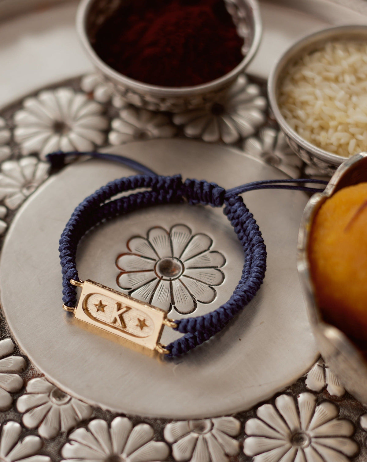 A blue bracelet with a geometric brick pattern, featuring a star-shaped charm, displayed on a decorative metal plate with floral designs and a small bowl of rice grains in the background.