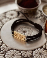 A jet black bracelet with a gold-tone brick bond design, displayed on a silver plate with floral patterns in the background.