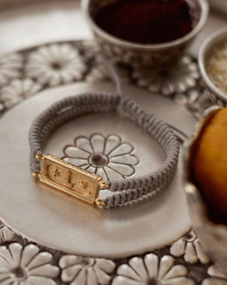 A smoke grey bracelet with a gold-colored initial 'L' on a flower-shaped charm, displayed on a silver plate with floral patterns in the background.